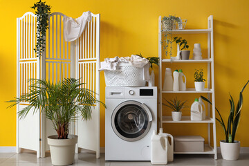 Modern washing machine with laundry and bottles of detergent near yellow wall