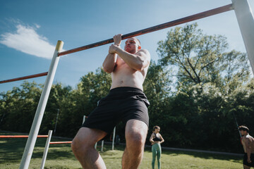 Athletic man performing pull-ups on bar in an urban park. Sunny day, exercise and fitness outdoors with other people in the background.