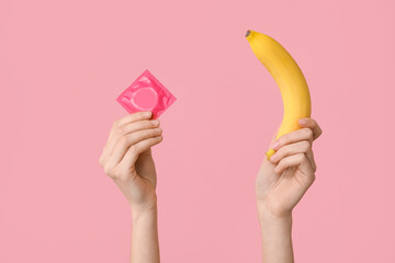 Female hands with banana and condom on pink background, closeup