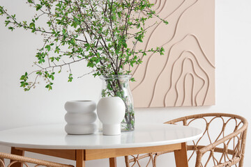 Table, armchairs and vase with blossoming branches near white wall in stylish dining room