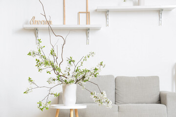 Vase with blooming branches on coffee table near cozy sofa in white living room