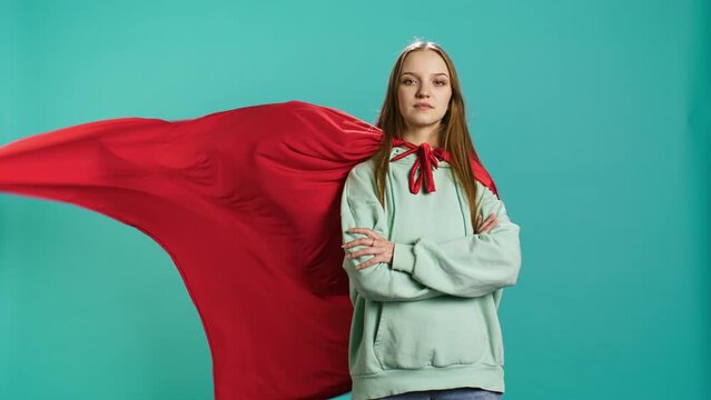 Happy young girl portraying superhero flying with cape fluttering in wind, isolated over studio background. Portrait of upbeat heroic woman posing as hero in costume, camera B