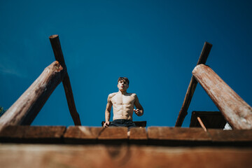 Athletic young man jumping from a wooden structure against a clear blue sky. His muscular physique...
