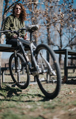 Fototapeta premium Outdoor portrait of a curly-haired young man leaning on a fence with his mountain bike, relaxed in a sunny park setting.