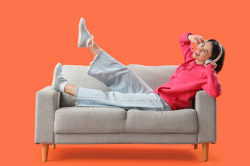 Happy young woman in headphones lying on sofa against orange background