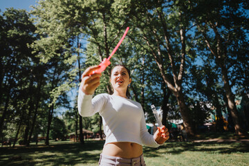 A young woman joyfully blows soap bubbles on a sunny day at the park, surrounded by lush green...