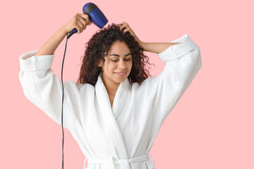 Beautiful young happy African-American woman blow drying her healthy curly hair on pink background