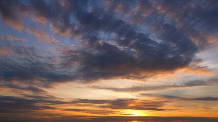 A dramatic sunset with the sky covered in dark, dense clouds. The setting sun casts a golden hue, creating a contrast between the dark clouds and the warm, glowing horizon. Sky background.
