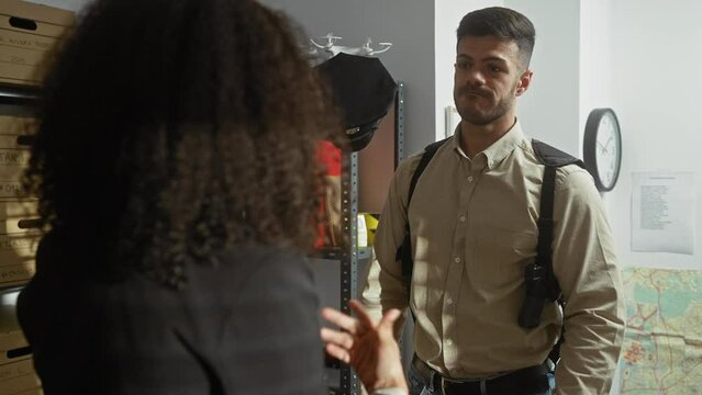 In an indoor setting, a woman and man, implied detectives, converse intently amidst investigation files and maps.