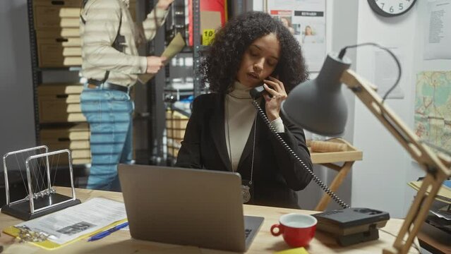 A focused woman investigator speaks on the phone, man examines files in a cluttered police office, portraying teamwork and crime investigation.