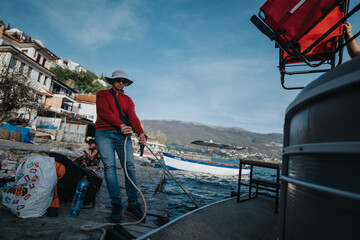 A man in a red shirt and white hat prepares a boat on a lakeside pier, surrounded by bags and a...