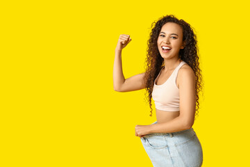 Happy young African-American woman in loose jeans showing muscles on yellow background. Weight loss concept