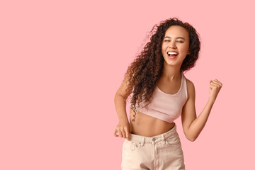 Happy young African-American woman in loose jeans showing yes gesture on pink background. Weight...