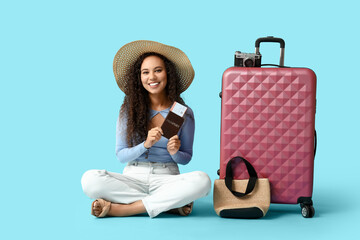 Happy young African-American woman with suitcase, passport and ticket sitting on blue background