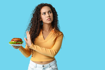 Young African-American woman showing stop gesture against burger on blue background. Diet concept