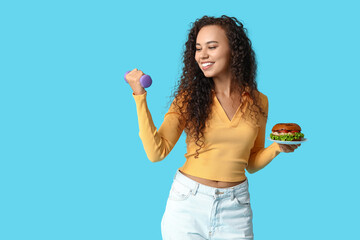 Happy young African-American woman with dumbbell and burger on blue background. Diet concept