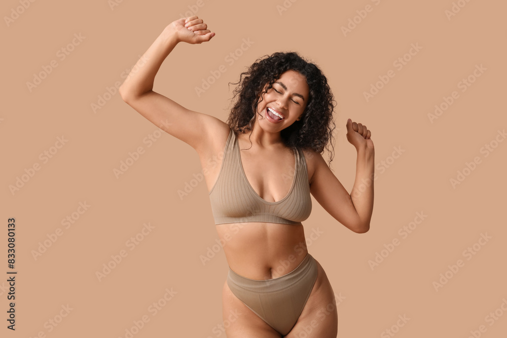 Wall mural Beautiful young happy African-American woman in underwear dancing on beige background