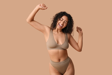 Beautiful young happy African-American woman in underwear dancing on beige background