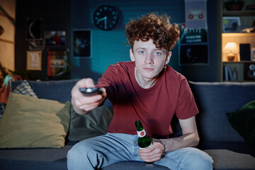 Medium shot of young Caucasian man holding bottle of beer and remote control turning on TV channel...