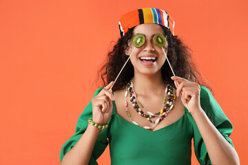Beautiful young happy African-American woman holding sticks with kiwi on orange background