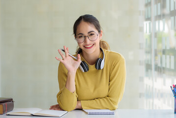 Confident woman with eyeglasses smiling while holding pen at desk, wearing headphone, relaxing in...