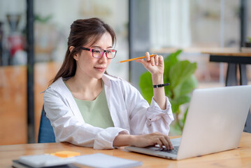 Young woman focused on her laptop in a modern office. She is holding a pencil, wearing glasses, and...