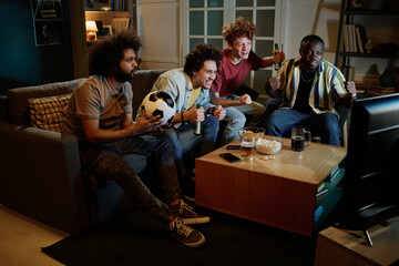 Group of joyful friends sitting in living room having fun watching soccer game match on TV late in...