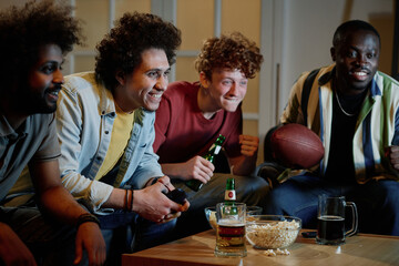 Medium shot of four multi-ethnic friends sitting on couch in living room having fun watching...