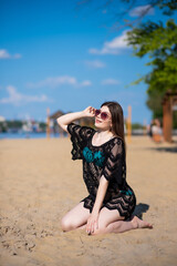 Beautiful, attractive, happy girl posing while sitting on a sandy beach on a sunny day wearing a blue swimsuit, sunglasses and a black lace beach tunic. Beachwear