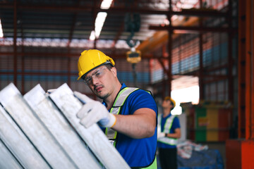 Caucasian male worker wearing a hard hat and safety glasses inspecting square pipe steel frame workpieces in iron factory.