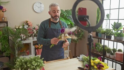 A bearded man arranges flowers in a florist shop with plants in the background, creating content for social media.