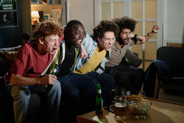 Multi-ethnic group of four young men sitting on couch in living room enjoying watching sports event...