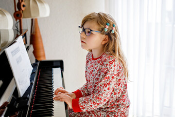 Little happy girl playing piano in living room. Cute preschool child with eye glasses having fun with learning to play music instrument.