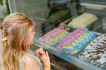 Happy preschool girl choosing and buying ice cream in outdoor stand cafe. Cute child looking at different sorts of icecream. Sweet summer dessert on family vacations. Summertime.