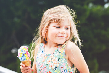 Happy preschool girl eating colorful ice cream in waffle cone on sunny summer day. Little toddler child eat icecream dessert. Sweet food on hot warm summertime days. Bright light, colorful ice-cream