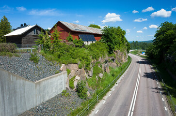 Road with a double white line "no crossing" stretching into the distance to forest, Wooden Scandinavian style farm buildings on a rock among green bushes and trees, Sunny summer day, Värmland, Sweden