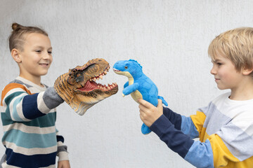 Two elementary school boys have fun playing role-playing games with dinosaur toys on a light background. Dinosaurs move in hands, focus on toys. Joyful communication, happy childhood