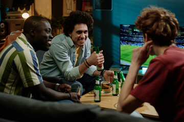 Ethnically diverse young men sitting in living room chatting while watching soccer match on TV late in evening