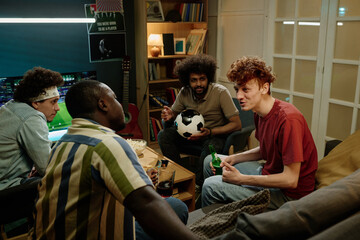 Multi-ethnic group of four young men sitting in living room discussing soccer match they watching...