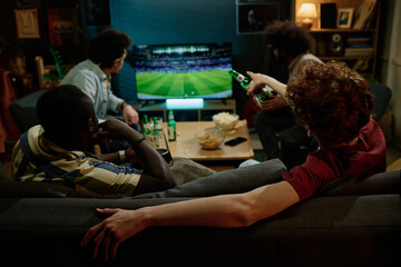Rear view of four young male friends spending evening indoors watching soccer match on TV and...
