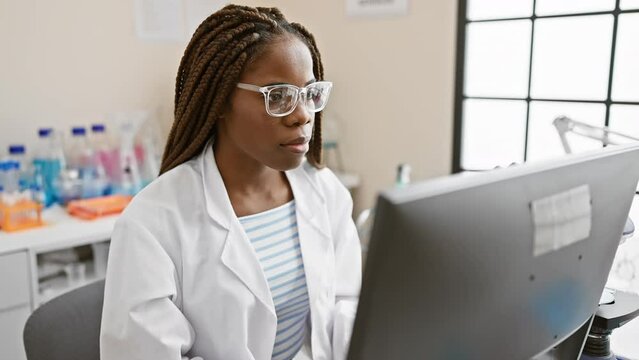 A focused black woman with braids wearing glasses and a white lab coat analyzing data on a computer in a laboratory setting