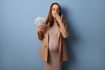 Shocked pregnant woman wearing beige jacket and dress holding big sum of euro banknotes standing...