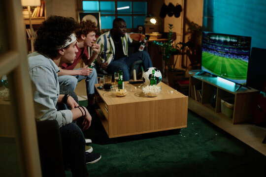 Group of multi-ethnic male friends sitting in living room watching soccer match on TV in evening, copy space
