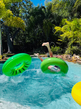 A boy flips upside down on a green raft in a swimming pool.