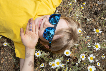 Portrait of a cute smiling boy in blue sunglasses lying among a chamomile field. Summer mood, childhood dreams, carefree joyful childhood. Hello summer, energy of nature. Earth Day. View from above