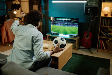 Unrecognizable biracial man holding ball sitting on sofa in living room watching soccer match on...