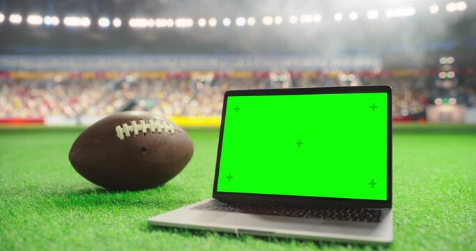 Laptop Computer with a Green Screen Mock Up Display Positioned Next to an American Football on a Stadium Pitch at Night. Template for Software, Streaming, Sports Betting, Multimedia Content