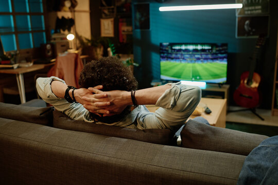 Rear view shot of unrecognizable man sitting relaxed in living room watching sports event on TV channel, copy space