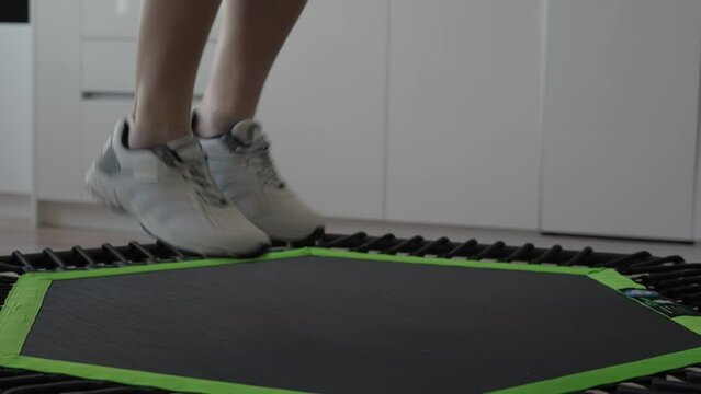 Close-up of a woman jumping on a sports trampoline at home, focusing on her legs and sneakers during an indoor workout.