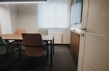 This image showcases a contemporary office meeting room with stylish wooden chairs and a matching table, set against a backdrop of a neatly positioned window blind and soft lighting.